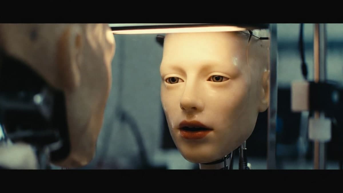 A humanoid robot with silicone skin practices facial expressions in front of a mirror in a laboratory setting
