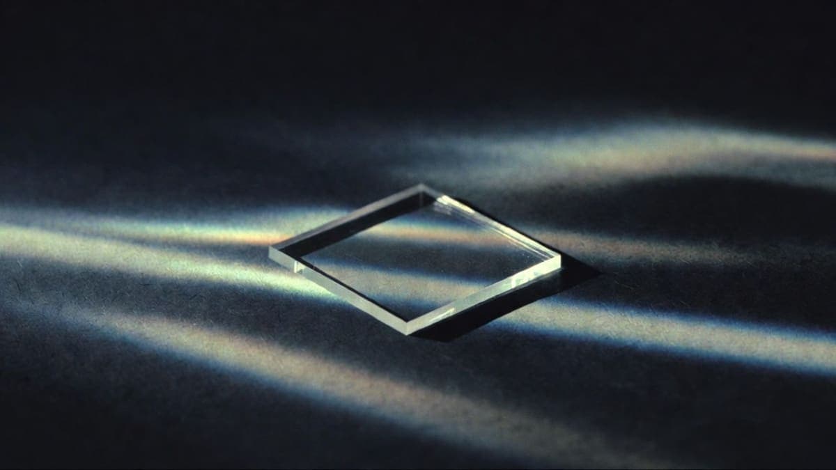 A small square of transparent borosilicate glass on a dark surface, refracting light through hundreds of laser-etched data layers
