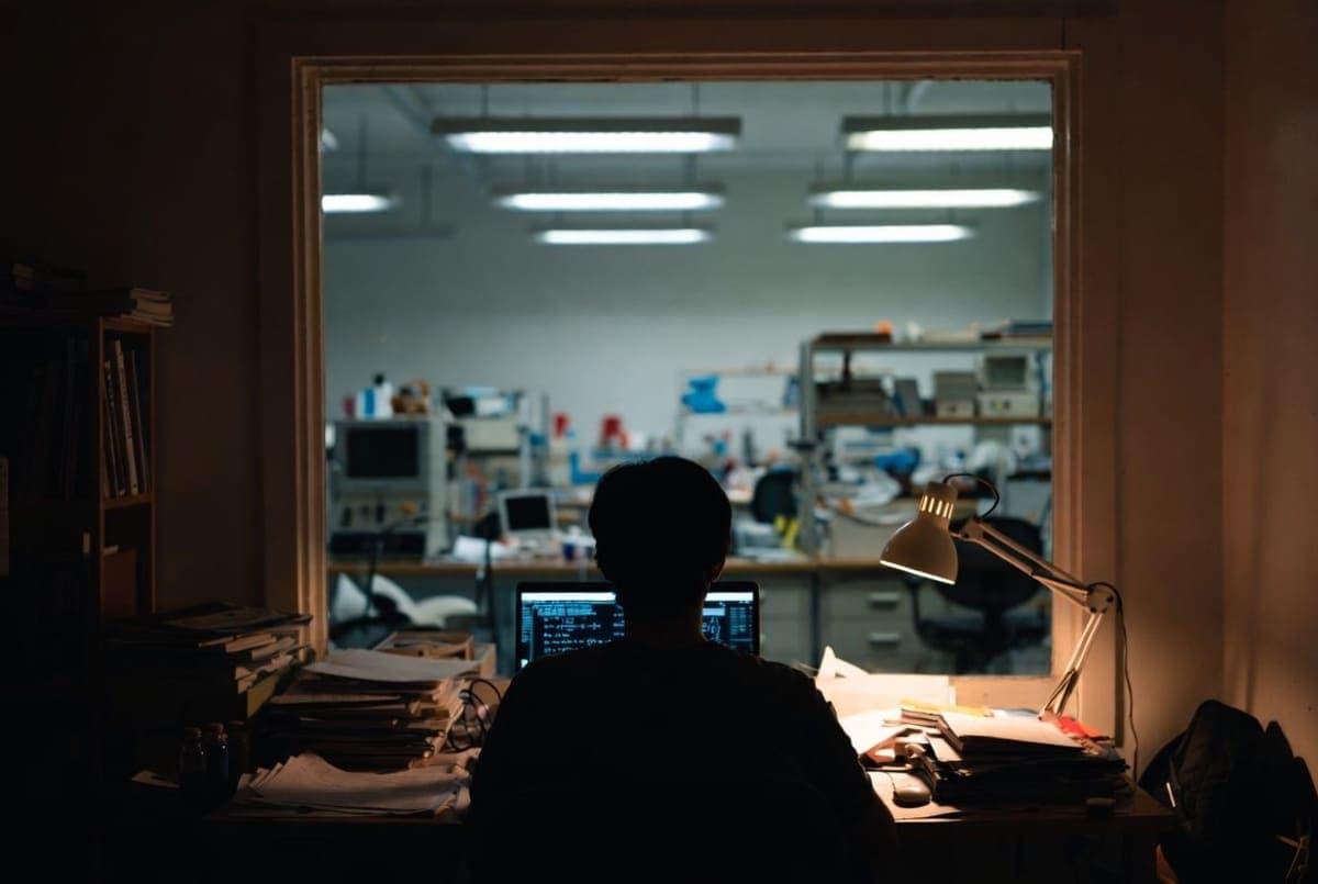 A researcher in a home office working on a laptop, with empty university laboratory equipment in the background