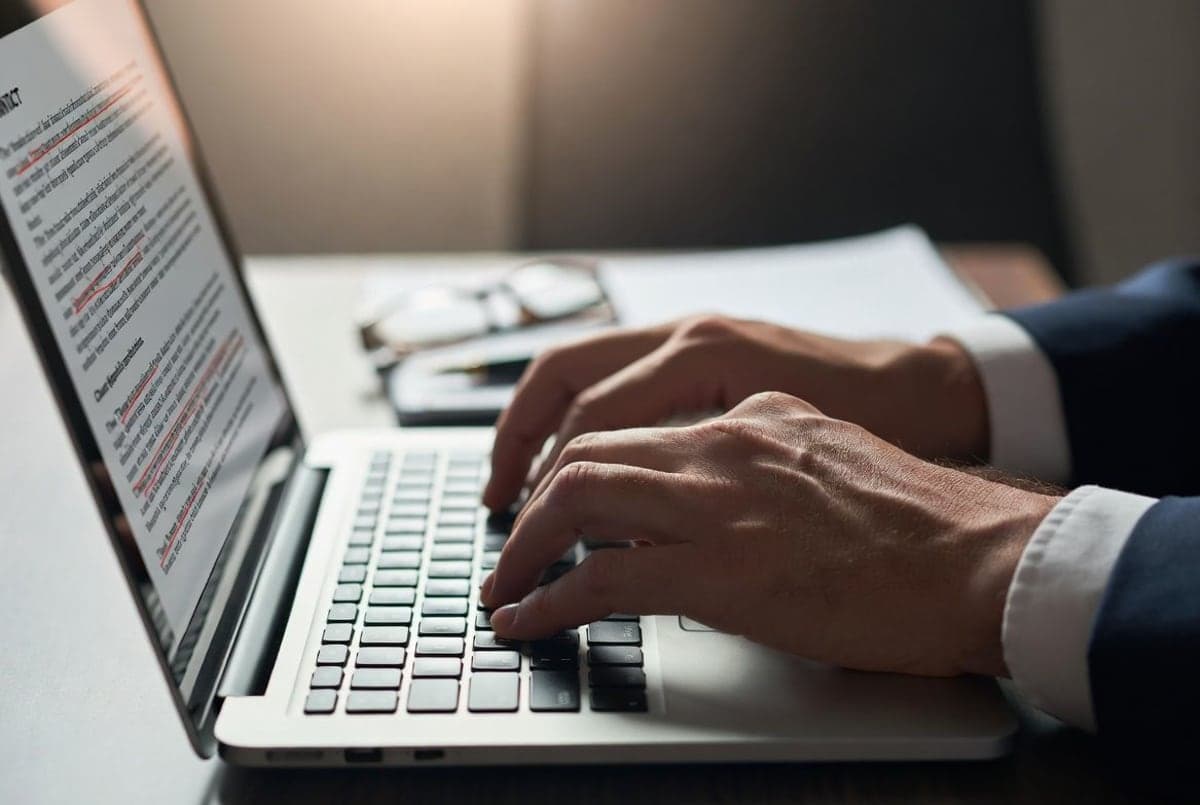 Lawyer's hands reviewing a contract on a laptop screen with red tracked-changes markings overlaying clauses