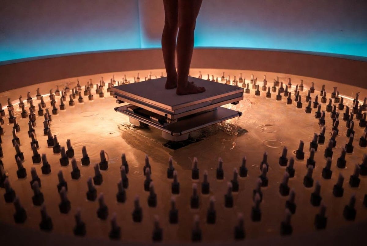 A person lowered into a shallow pool surrounded by a ring of ultrasound transducers in a clinical wellness setting