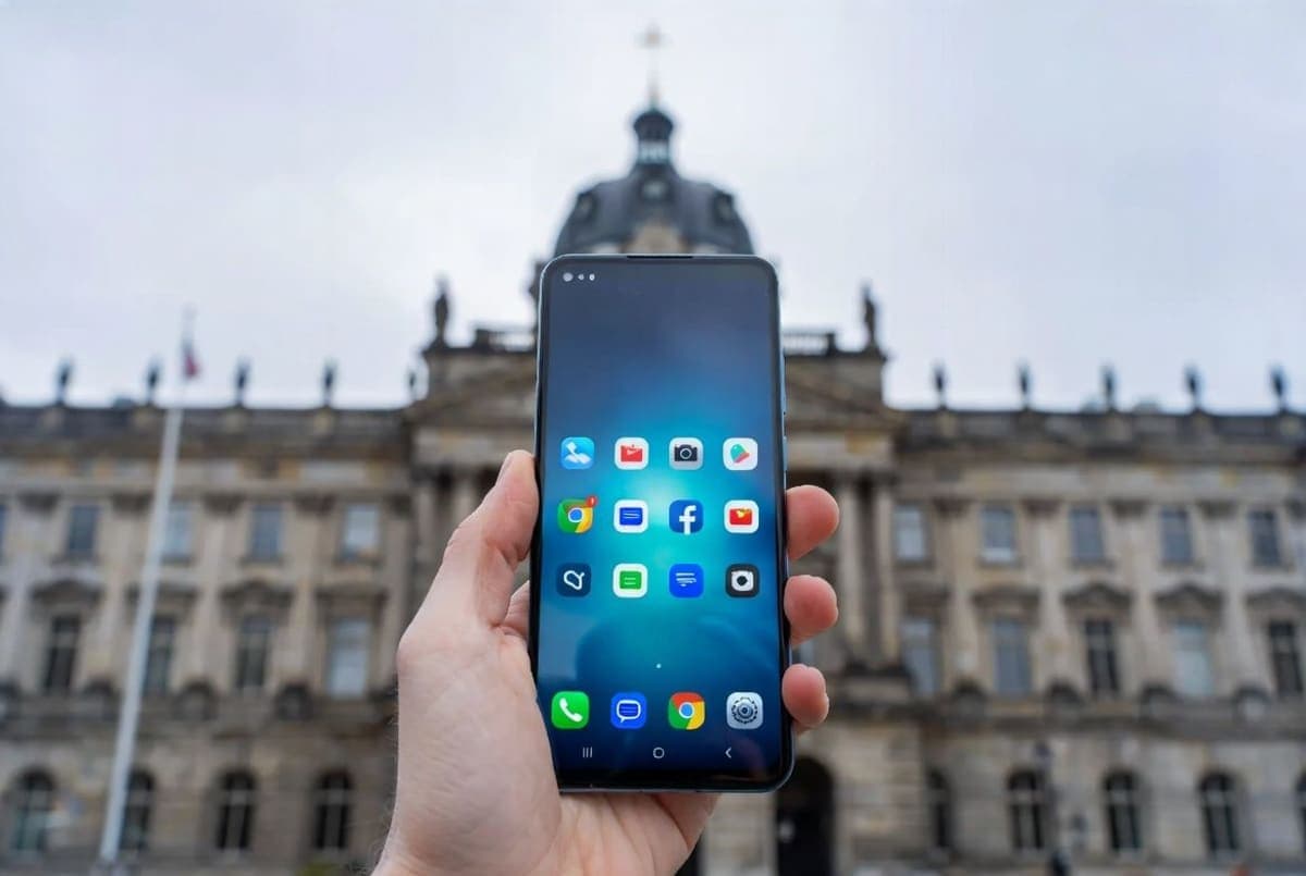Android smartphone displaying app icons, held in a hand against a blurred European city backdrop